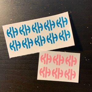 May include: Two sheets of monogram stickers. One sheet has blue stickers, and the other has pink stickers. Each sticker features a stylized monogram design. The stickers are on a white background.