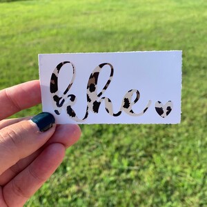 May include: A white sticker with the word "she" written in a cursive font with a leopard print pattern. A small heart with a leopard print pattern is to the right of the word.