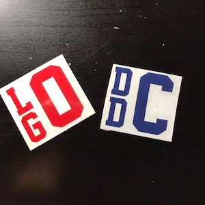 May include: Two square white vinyl decals featuring bold block lettering. The left decal displays the red letters LG and O, while the right decal features the blue letters D, D, and C arranged in a vertical and horizontal layout.
