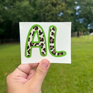 May include: A rectangular white decal featuring the letters AL in a bold, rounded font. The letters have a brown and black leopard print pattern and are outlined with a thick, vibrant lime green border against a plain white background.