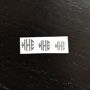 May include: Three gray monogram stickers with the letters KHE inside an oval shape, arranged in a row on a white rectangular sticker. The stickers are on a dark wood surface.