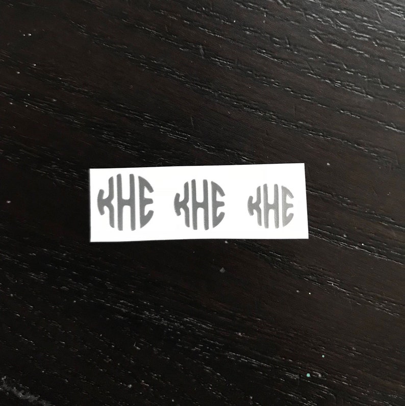 May include: Three gray monogram stickers with the letters KHE inside an oval shape, arranged in a row on a white rectangular sticker. The stickers are on a dark wood surface.