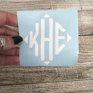 May include: A white vinyl decal featuring a monogram with the letters K, H, and E arranged in a vertical diamond shape. The design is centered on a light blue backing sheet, with small white triangles pointing outward at the top, bottom, left, and right.