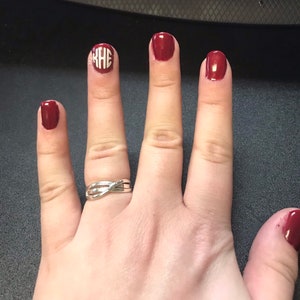 May include: Hand with burgundy nail polish on four fingers and a silver ring. One nail features a white monogram design. The nails are painted with a glossy finish. The ring has a twisted design.