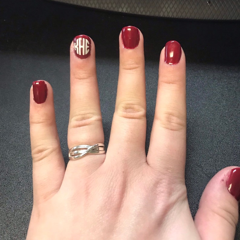May include: Hand with burgundy nail polish on four fingers and a silver ring. One nail features a white monogram design. The nails are painted with a glossy finish. The ring has a twisted design.