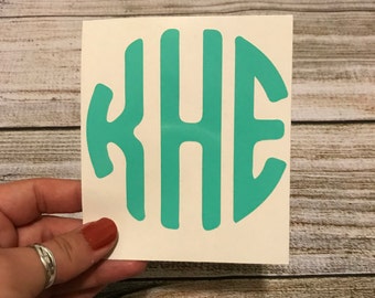 May include: A teal vinyl decal featuring a circular monogram design with the letters K, H, and E in a stylized, bold font. The letters are interconnected to form a circular shape, presented on a white backing sheet for application.