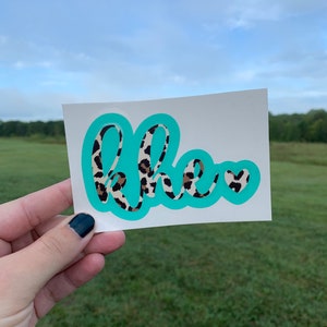 May include: A rectangular white sticker featuring the lowercase letters "khe" in a cursive font with a brown and black leopard print pattern. The letters are outlined by a thick, solid turquoise border, followed by a small turquoise heart.