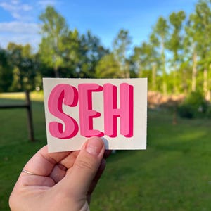 May include: A rectangular white sticker featuring the letters SEH in a bold, bubble-style font. The letters are colored in a vibrant pink with a darker pink shadow effect, creating a layered appearance on the white background surface.