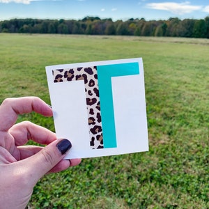 May include: A square white decal featuring a large capital letter T. The left half of the letter is covered in a brown and black leopard print pattern, while the right half is a solid, vibrant teal color. The decal is presented on a white backing.