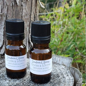 May include: Two amber glass bottles with black caps and white labels. The labels read "Scentered Aroma Center & Soothe Diffuser Blend" with the text "with fir and sweet orange essential oils" on the bottom.