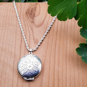 May include: A silver chain necklace with a round silver locket pendant. The locket features a detailed floral design.