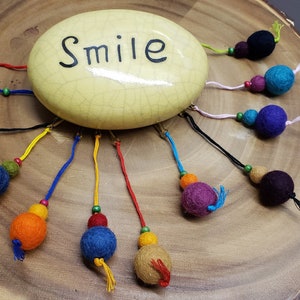 May include: A white ceramic stone with the word "Smile" written on it. The stone has a variety of colourful felt balls attached to it with strings.