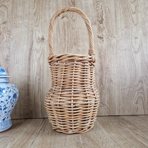 May include: A tall, light brown woven wicker basket featuring a rounded base that tapers towards the middle before widening slightly at the top, finished with a single arched handle attached to the rim, showcasing a natural textured weave pattern.