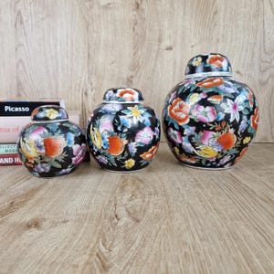 May include: Three black ceramic ginger jars with lids, adorned with a colourful floral pattern in pink, orange, yellow, and blue. The jars are of varying sizes and have a glossy finish. They are set on a wooden surface.