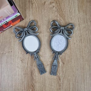 May include: Two matching oval picture frames in a metallic silver finish, each featuring a decorative bow at the top and a tassel detail at the bottom. The frames have a beaded edge design, with one containing a plain white insert and the other a floral pattern.