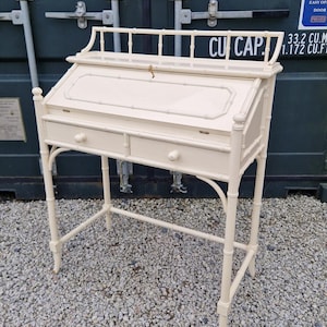 May include: A cream-coloured bureau desk featuring a faux bamboo frame, a drop-front writing surface with a small gold key, two horizontal drawers with round knobs, and a decorative gallery rail along the top edge of the desk surface.