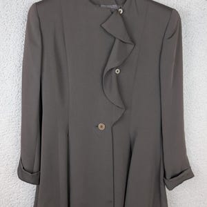 May include: A dark grey blazer featuring a high neckline, long sleeves with folded cuffs, and a vertical ruffle detail along the front closure. The garment is secured with three visible buttons and is displayed on a wooden hanger.