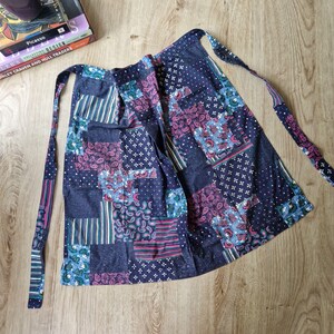 May include: A patchwork apron in blue, pink, and white. The apron has a pocket and long ties. The design incorporates various patterns, including paisley and stripes. The fabric appears to be cotton.