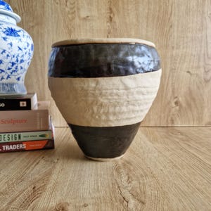 May include: A handcrafted ceramic vase with a distinctive design. The vase has a black top and bottom with a textured, cream-coloured band in the middle. The vase has a wide opening and a slightly tapered shape. The vase is a decorative item.
