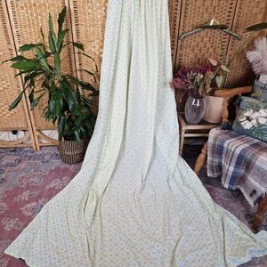 May include: A long, pale green curtain with a floral pattern hangs from a wooden hanger. The curtain is draped, creating a flowing effect. The background includes a woven room divider, plants, and a patterned rug.