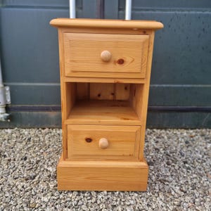 May include: A pine bedside cabinet with two drawers and an open shelf. The drawers have round wooden knobs. The cabinet is made of light-coloured wood and has a simple, classic design.