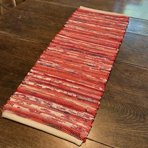 May include: A rectangular, handwoven rug with a vibrant red and white color scheme. The rug features a striped pattern with varying shades of red, pink, and white. The ends are finished with a cream-colored border. The rug is placed on a wooden surface.