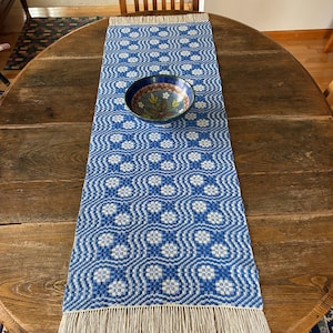 May include: A blue and white patterned table runner with a fringed edge, displayed on a wooden table. A decorative bowl with a floral design sits atop the runner. The runner's pattern features a repeating geometric design.