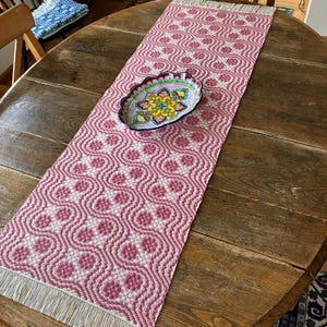 May include: A woven pink and white patterned table runner with a fringed edge, displayed on a round wooden table. A decorative oval dish with a colorful floral design sits on the runner. The runner's pattern is a repeating geometric design.