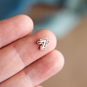 Puede incluir: Un pequeño dije dorado con forma del número siete. El dije tiene un contorno rectangular con el número siete en el interior. El dije tiene dos pequeños bucles a cada lado. El dije se sostiene entre dos dedos.