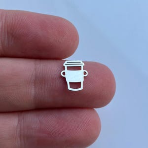 Puede incluir: Un amuleto de plata con forma de taza de café con tapa. El amuleto es pequeño y detallado.