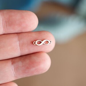 Könnte beinhalten: Ein kleiner, goldfarbener Unendlichkeitsanhänger. Der Anhänger wird zwischen den Fingern gehalten, was sein filigranes Design hervorhebt. Das Unendlichkeitssymbol ist eine durchgehende Schleife, die oft in Schmuck verwendet wird, um Ewigkeit oder unendliche Liebe darzustellen.