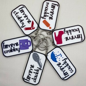 May include: Six white rectangular keychains with black borders and different colored illustrations and text. The keychains read "femme stabby", "femme crabby", "femme dacky fruity", "femme happy mackey", "femme happy socks", and "femme happy baddi".