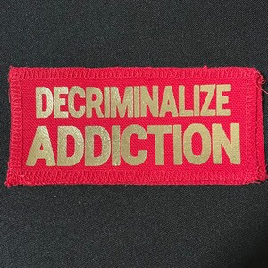 May include: Red fabric patch with gold lettering that reads "DECRIMINALIZE ADDICTION".