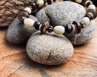 Puede incluir: Un brazalete de cuentas hecho con materiales naturales. Incluye cuentas marrones, beige y blanquecinas, con algunos detalles metálicos. El brazalete se exhibe sobre una superficie de madera con piedras y una cesta tejida en el fondo. Circunferencia aproximada de 19 cm.