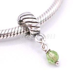 Puede incluir: Un charm de plata con un diseño de nudo y una cuenta verde colgante. El charm está unido a una pulsera de plata. La cuenta es de color verde claro y tiene una superficie facetada. El charm es probablemente de plata de ley.