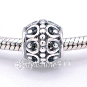 May include: A silver charm bead featuring an intricate openwork pattern of curved lines and raised dots, threaded onto a matching silver snake chain bracelet. The bead has a polished finish with darkened recessed areas to highlight the design details.