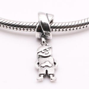 Puede incluir: Un charm de plata con forma de niño, con gorro y pantalones cortos, colgado de una pulsera de plata. El charm está detallado con rasgos faciales y ropa. La pulsera es una cadena de serpiente de plata.