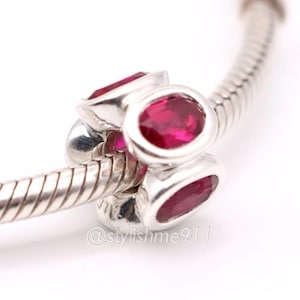 May include: A silver charm with three oval, deep red gemstones. The charm is on a silver bracelet. The gemstones are set in a silver bezel. The charm is a bead style. The background is white.