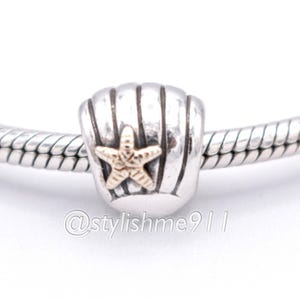 Pode incluir: Um berloque de prata com um design de concha e uma estrela do mar dourada no centro. O berloque está em uma pulseira de prata. O berloque é uma joia.