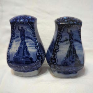 May include: Pair of blue and white ceramic salt and pepper shakers with images of the Pilgrim Monument in Provincetown, Massachusetts. The shakers are decorated with floral designs and the words "Pilgrim Monument" and "A.D. 1910".