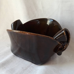 May include: A dark brown ceramic bowl with a glossy finish. The bowl has a rectangular shape with curved sides and a handle on each end. The bowl is sitting on a white surface.