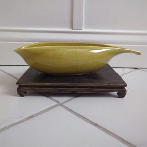 May include: A yellow ceramic bowl with a leaf-like shape and a spout. The bowl is sitting on a dark wooden stand.