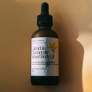 May include: A dark brown glass bottle of Calendula Chamomile Infused Body Oil with a black dropper. The label features text and a yellow flower illustration. The bottle is on a light brown surface.