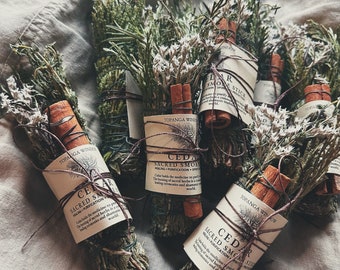 Puede incluir: Un conjunto de cuatro manojos de hierbas y flores secas atados con cuerda. Cada manojo está etiquetado con "Cedar Sacred Smoke" y "Topanga Winds". Los manojos están atados con palitos de canela y están listos para ser quemados para la limpieza con humo.