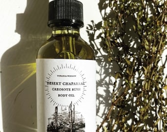 May include: A glass bottle of Desert Chaparral Creosote Bush Body Oil with a black dropper top and a white label with black text and a desert scene illustration. The label reads "Topanga Window Desert Chaparral Creosote Bush Body Oil".