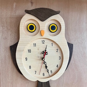 Puede incluir: Reloj de madera con forma de b&uacute;ho con una cara marr&oacute;n y ojos amarillos. La esfera del reloj tiene n&uacute;meros negros y una aguja de hora roja y una aguja de minutos negra.