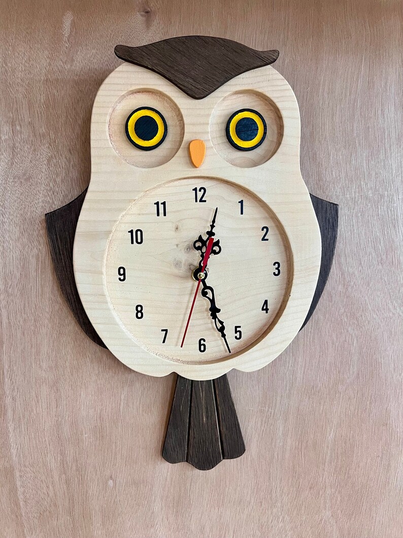 Puede incluir: Reloj de madera con forma de b&uacute;ho con una cara marr&oacute;n y ojos amarillos. La esfera del reloj tiene n&uacute;meros negros y una aguja de hora roja y una aguja de minutos negra.