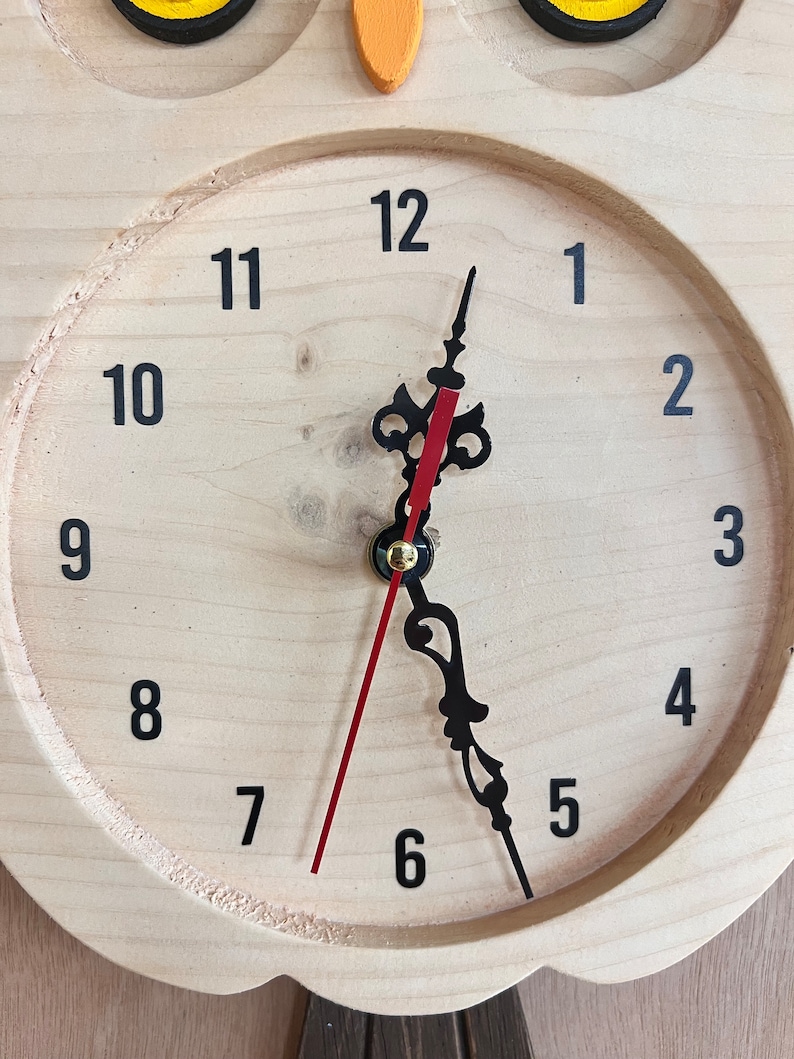 Puede incluir: Un reloj de madera con forma de b&uacute;ho con n&uacute;meros y manecillas negros. La esfera del reloj es de color marr&oacute;n claro con una manecilla de hora roja y una manecilla de minutos negra. Los ojos del b&uacute;ho son amarillos y el pico es naranja.