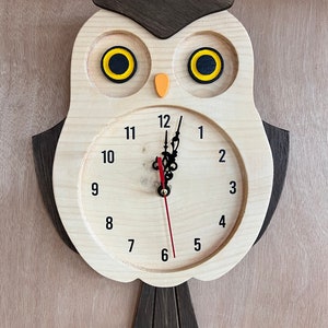 Puede incluir: Reloj de madera con forma de b&uacute;ho con una cara de b&uacute;ho marr&oacute;n y ojos amarillos. La esfera del reloj es blanca con n&uacute;meros negros del 1 al 12 y agujas negras.