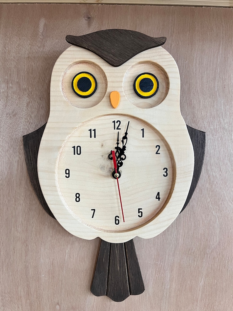 Puede incluir: Un reloj de madera con forma de b&uacute;ho con una aguja de hora roja y una aguja de minutos negra. Los ojos del b&uacute;ho son amarillos con pupilas negras. La esfera del reloj tiene n&uacute;meros negros del 1 al 12.